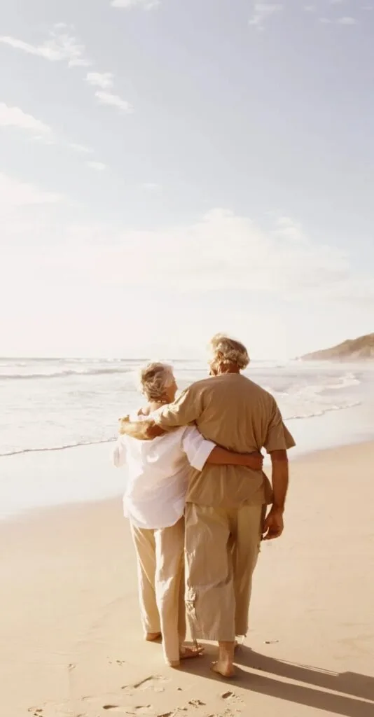 An elderly couple walking on the beach arm in arm. 