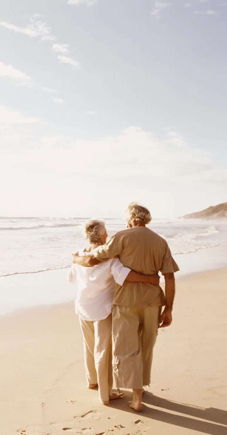 An elderly couple walking on the beach arm in arm. 